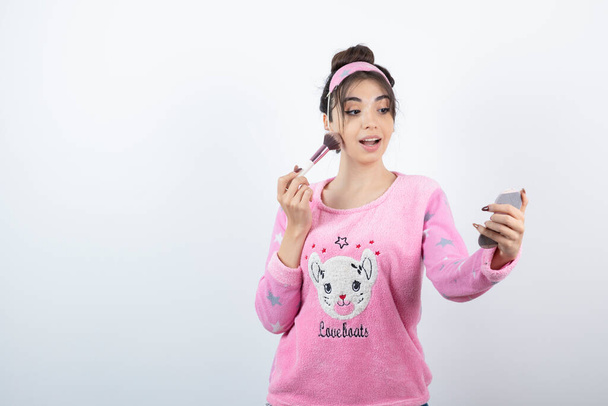 Young woman in pink pajama applying powder while looking at mirror. High quality photo - Photo, Image