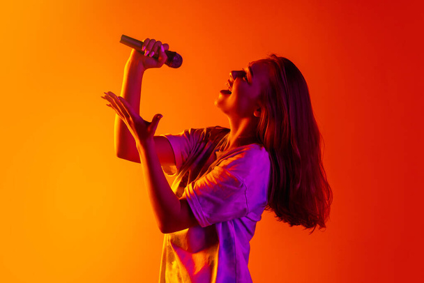 Portrait recadré latéral de jeune belle fille caucasienne chantant avec microphone sur fond rouge jaune dégradé - Photo, image