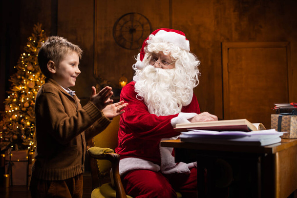 Santa Claus and little boy. Cheerful Santa is working while sitting at the table. Fireplace and Christmas Tree in the background. Christmas concept. - Foto, Imagen