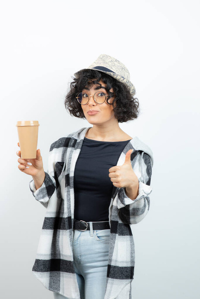 Imagen de la hermosa morena hipster sosteniendo una taza de café y mostrando los pulgares hacia arriba. Foto de alta calidad - Foto, imagen
