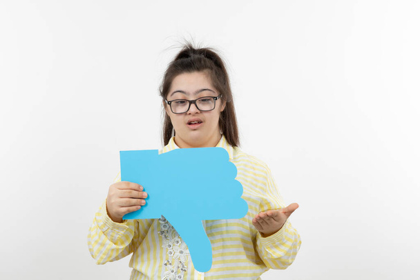 menina com síndrome de down posando com papel de corte em forma de mão azul, polegar para baixo - Foto, Imagem