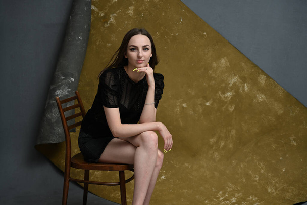 A full-length photo of a young brunette girl sits on a chair in the studio on a gray-golden background - Foto, immagini