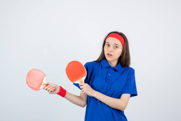 κορίτσι σε μπλε t-shirt, κόκκινο περικάρπιο και κορδέλα ποζάρουν με ρακέτες τένις - Φωτογραφία, εικόνα