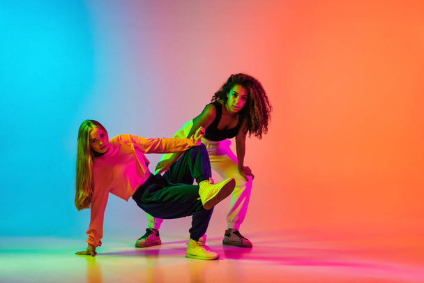 Rhytm. Two stylish modern girls dancing hip-hop on gradient blue orange background in neon - Фото, изображение