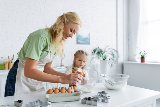 vrouw breken ei in kom tijdens het koken met dochter in de keuken - Foto, afbeelding