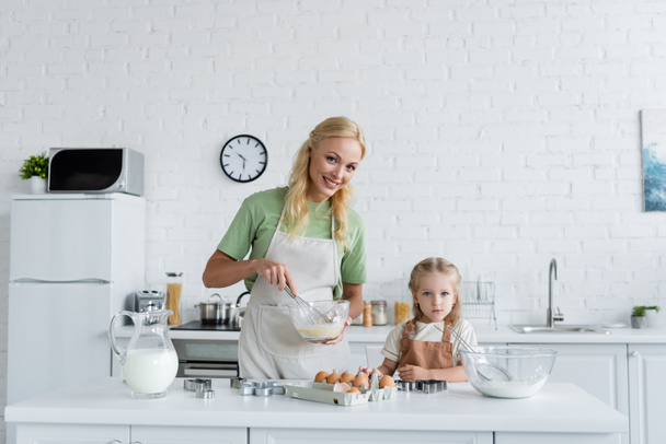 веселая женщина смешивает ингредиенты с венчиком рядом с маленькой дочерью на кухне - Фото, изображение