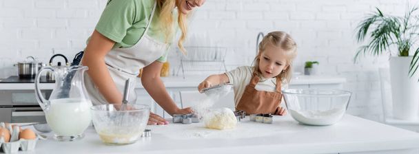 fille tamiser la farine sur la pâte près de la mère souriante et les ingrédients sur la table, bannière - Photo, image