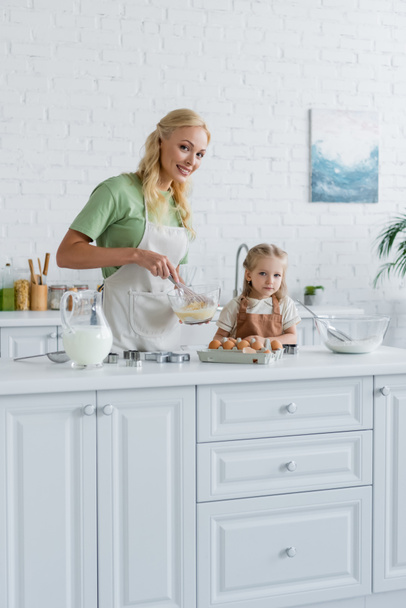 счастливая мама смотрит в камеру, смешивая ингредиенты в миске рядом с дочерью - Фото, изображение