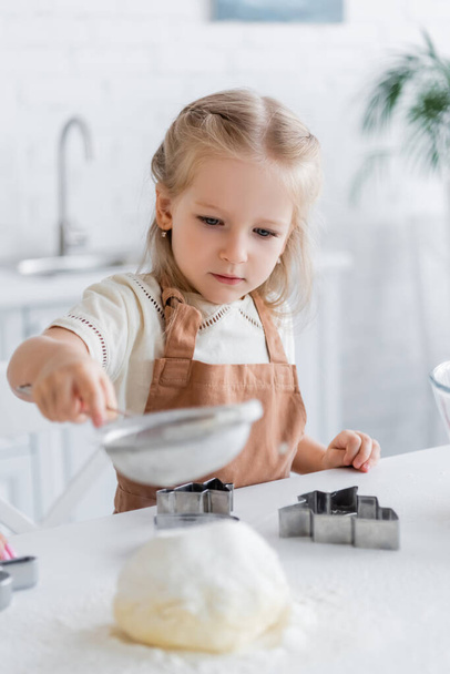 child in apron sifting flour near cookie cutters, blurred foreground - Foto, Imagen