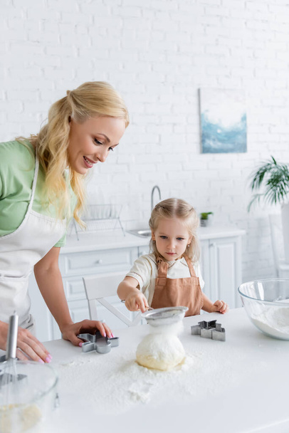 το παιδί κοσκινίζει αλεύρι σε ακατέργαστη ζύμη ενώ βοηθά τη μαμά στην κουζίνα - Φωτογραφία, εικόνα