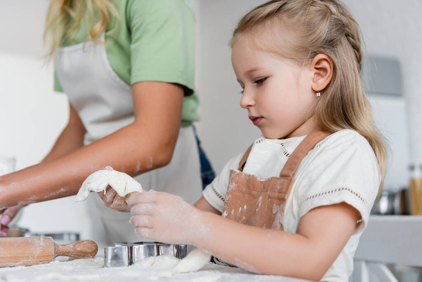 παιδί κρατώντας ζύμη κοντά σε κόφτες μπισκότων και θολή μαμά στην κουζίνα - Φωτογραφία, εικόνα