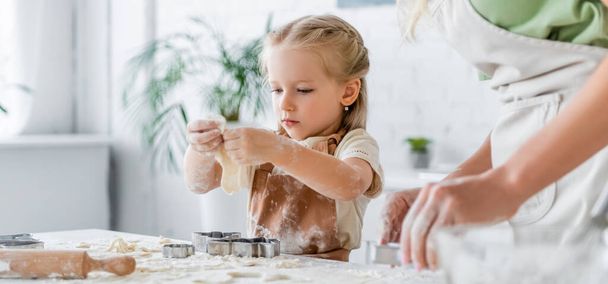 girl in apron near blurred mother cutting raw dough with cookie cutter, banner - Foto, Imagem