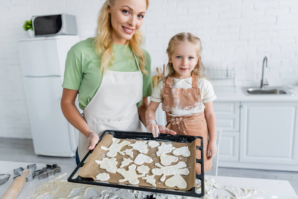 gelukkig moeder en dochter houden bakplaat met rauwe multi-vormige koekjes - Foto, afbeelding