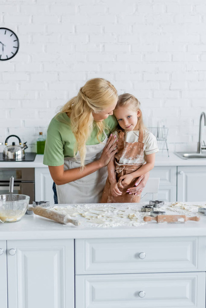 γυναίκα αγκαλιάζει την κόρη κοντά στο τραπέζι της κουζίνας με αλεύρι και μαγειρικά σκεύη - Φωτογραφία, εικόνα