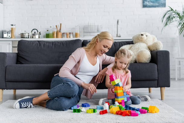 girl making tower of building blocks near mom and teddy bear on couch - Photo, Image