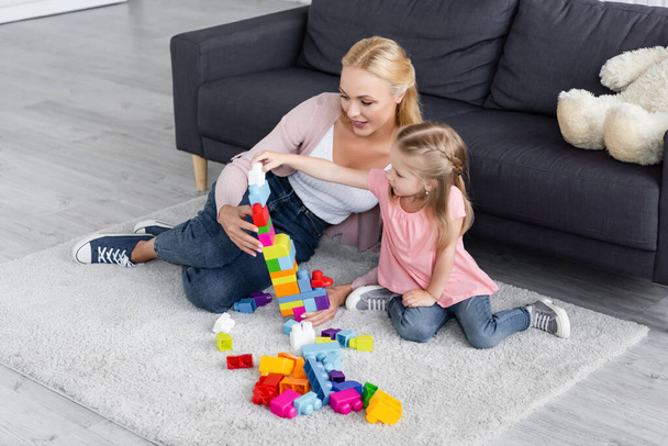 child making tower of building blocks near mom on floor at home - Фото, изображение