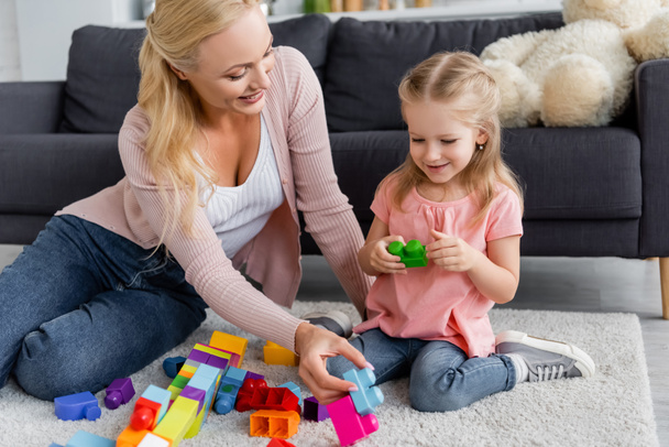 happy woman with smiling daughter playing with multicolored cubes on floor at home - 写真・画像
