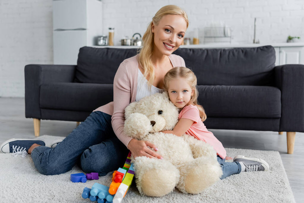 mother and daughter embracing on floor near teddy bear and colorful building blocks - Photo, Image