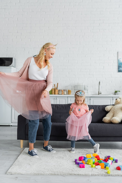 happy woman and girl in tulle skirts and toy crowns dancing in kitchen - Photo, Image
