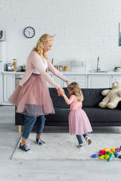 happy woman and daughter in costumes of princesses dancing in kitchen - Photo, Image