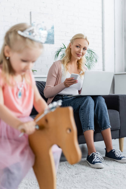 woman with laptop and mobile phone looking at blurred daughter riding rocking horse - Photo, Image
