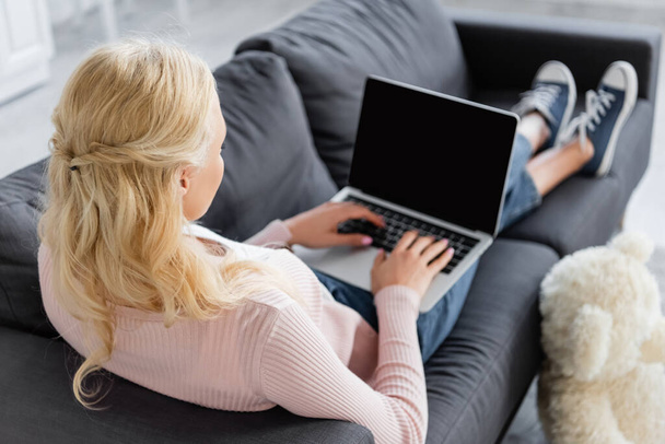 mulher loira sentada no sofá e digitando no laptop com tela em branco - Foto, Imagem