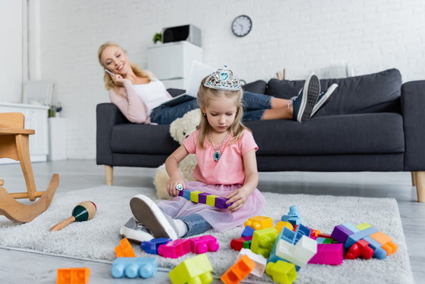 blurred woman talking on smartphone near daughter playing with building blocks on floor - Fotografie, Obrázek