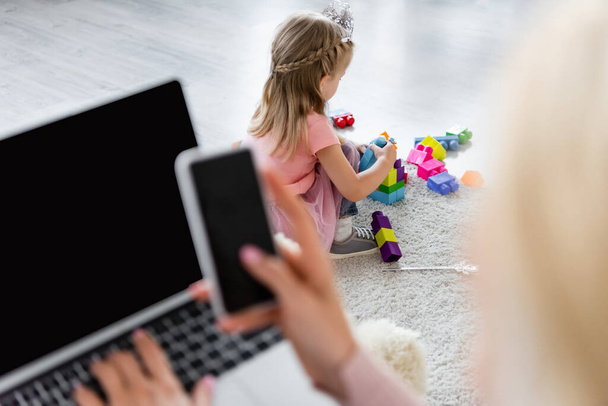 bambino che gioca sul pavimento con mattoni vicino alla mamma con gadget in primo piano sfocati - Foto, immagini