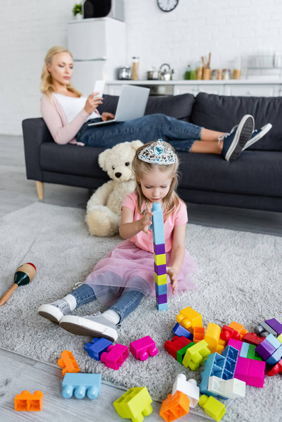 chica en juguete corona jugando en el suelo con bloques de construcción cerca de mamá con gadgets sobre fondo borroso - Foto, Imagen