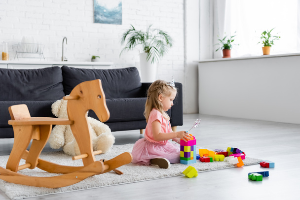 girl in princess costume playing with magic wand and building blocks near rocking horse - Photo, Image