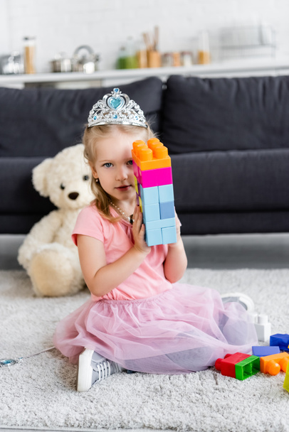 girl in toy crown showing tower of multicolored building blocks while sitting on floor - Foto, imagen