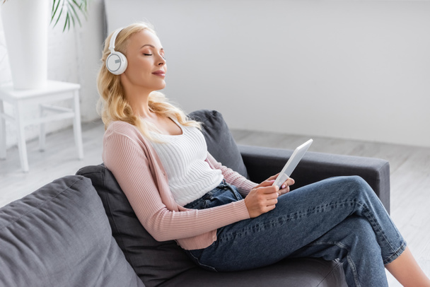 mujer rubia con tableta digital escuchando música mientras está sentada en el sofá con los ojos cerrados - Foto, Imagen