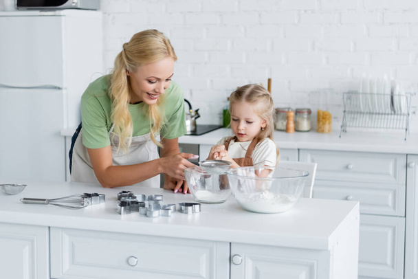 girl sifting flour with sieve into bowl near happy mother - Fotó, kép