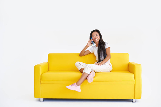Smiling asian woman talking on cell phone sitting on yellow couch - Foto, immagini