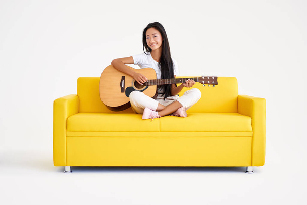 Smiling pretty female playing guitar sitting on yellow sofa on white background - Foto, afbeelding