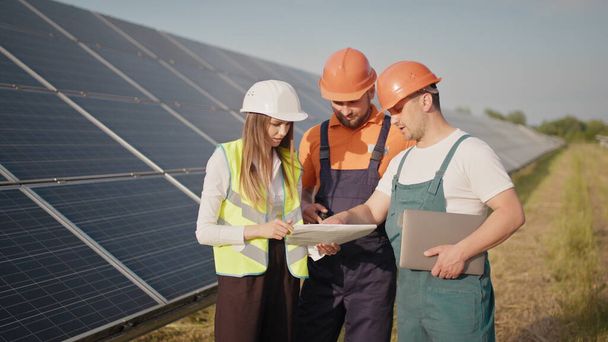 Inženýři používají kresby na solární farmě ve speciálních uniformách stojící rozhlédnout diskutovat o instalaci slunečných baterií elektřina zpomalení životního prostředí. Setkání s investorem - Fotografie, Obrázek