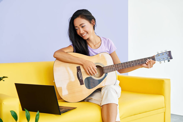 Smiling pretty female learning to play guitar watches a video lesson online using laptop - Fotoğraf, Görsel
