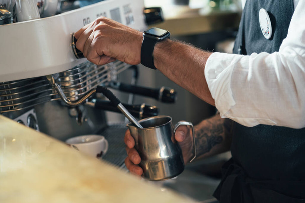 Ένας ανώνυμος μπάρμαν φτιάχνει ένα καπουτσίνο σε ένα καφέ. Κοντινό πλάνο φωτογραφία ενός άνδρα χέρια με τατουάζ χρησιμοποιώντας μια καφετιέρα για να προετοιμάσει ένα φλιτζάνι καφέ σε ένα καφέ. - Φωτογραφία, εικόνα