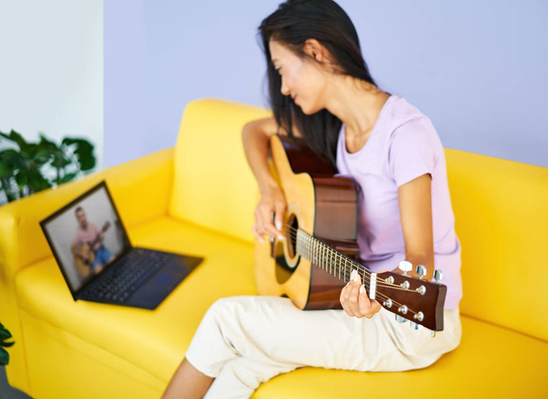 Smiling pretty female learning to play guitar watches a video lesson online using laptop - Zdjęcie, obraz