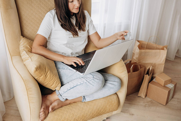 Женщина держит кредитную банковскую карту и использует ноутбук для покупок в Интернете дома. Улыбающаяся девушка сидит в кресле и использует ноутбук для электронной коммерции. Селективный фокус - Фото, изображение