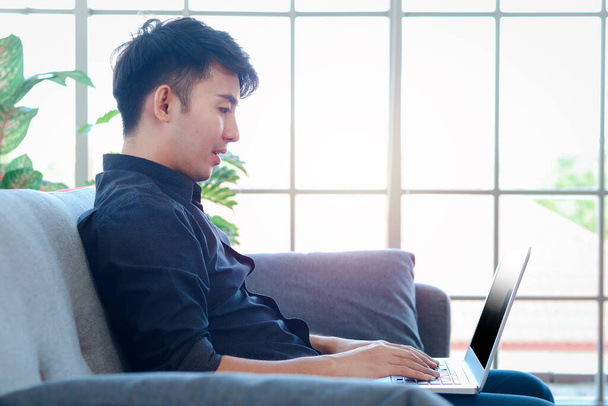 Азиатский молодой бизнесмен, печатающий на клавиатуре ноутбук, сидя на диване в своей квартире, фрилансер, работающий онлайн в гостиной.  - Фото, изображение