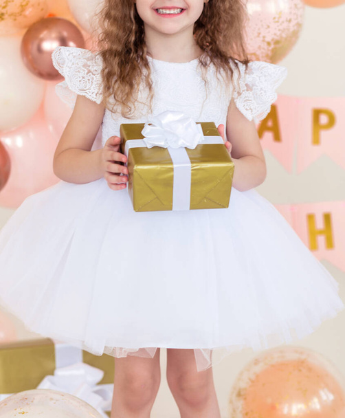 Маленька дівчинка в білій пухнастій сукні позує з подарунковою коробкою в руках. Фотографія
 - Фото, зображення