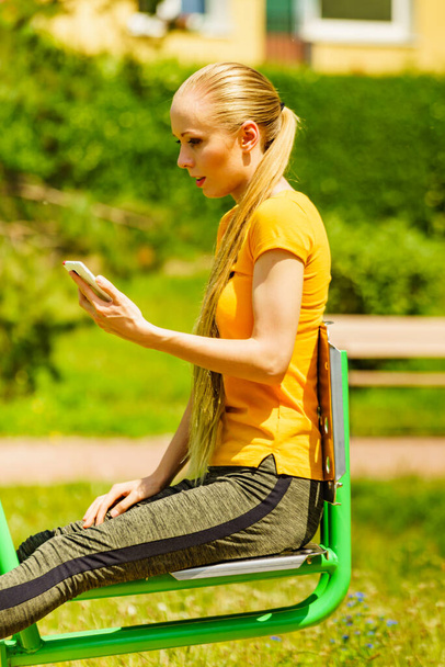 Young woman working out in outdoor gym. Girl holding mobile phone while doing exercises on street machine. - Foto, Bild