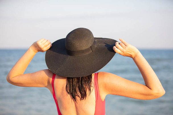Una donna con un cappello a tesa larga e un costume da bagno guarda il mare. - Foto, immagini