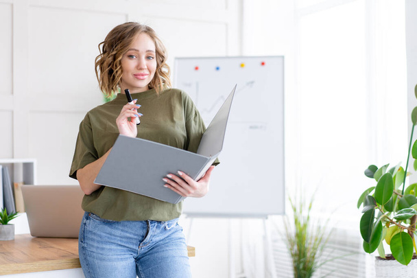 Businesswoman holding folder for paper write marker thinking dreaming standing near office table. Business person femal ypoung adult caucasian on modern white interior  - Foto, afbeelding