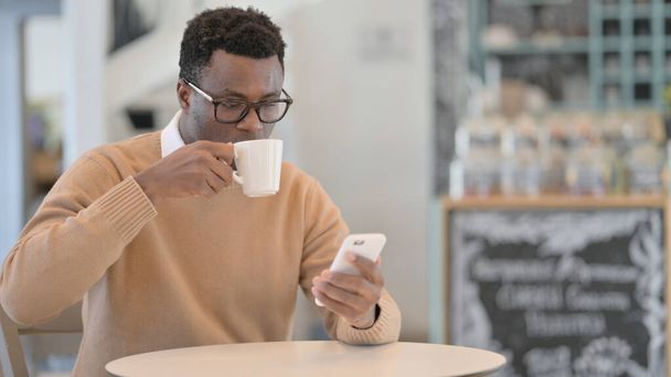 African Man using Smartphone and Drinking Coffee in Cafe - Фото, зображення