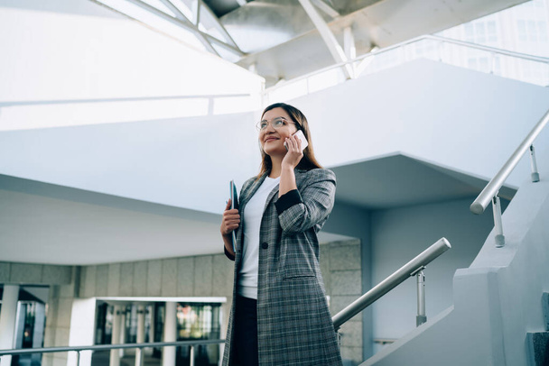 Smiling hipster girl in trendy outfit and eyewear making mobile phone call standing on urban architecture stairs, positive caucasian woman satisfied with roaming connection talking on smartphone - Foto, Bild