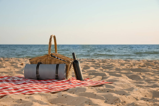 Blanket with picnic basket and bottle of wine on sandy beach near sea, space for text - Foto, afbeelding