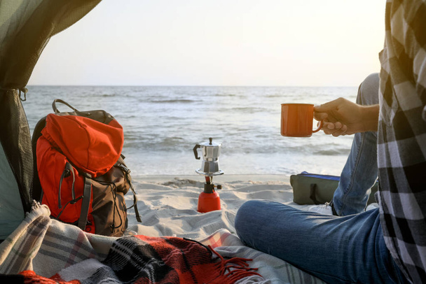 キャンプ場のテントの中で熱い飲み物のカップを持つ男、クローズアップ - 写真・画像