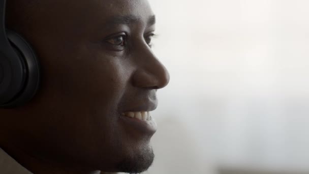 Африканский человек слушает музыку онлайн в наушниках в помещении, приседает, смотрит сбоку - Кадры, видео